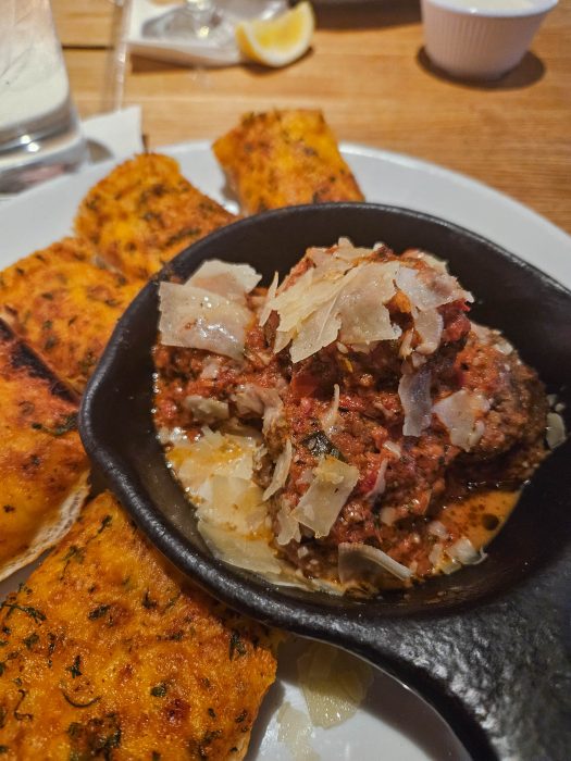 A close-up of a skillet with meatballs topped with tomato sauce and shaved cheese, served with slices of seasoned bread on a white plate.