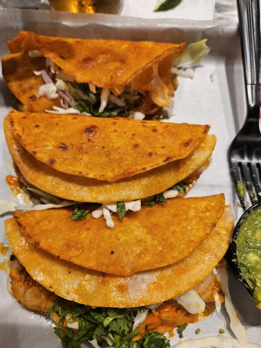 Three folded tacos with orange crispy shells, filled with shredded cabbage, herbs, and other ingredients, placed on a tray next to a plastic fork and a small container of green sauce.