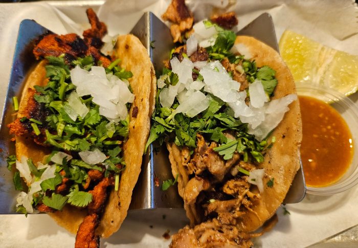 Two tacos in a metal holder, topped with chopped onions and cilantro, served with lime wedges and a side of salsa.