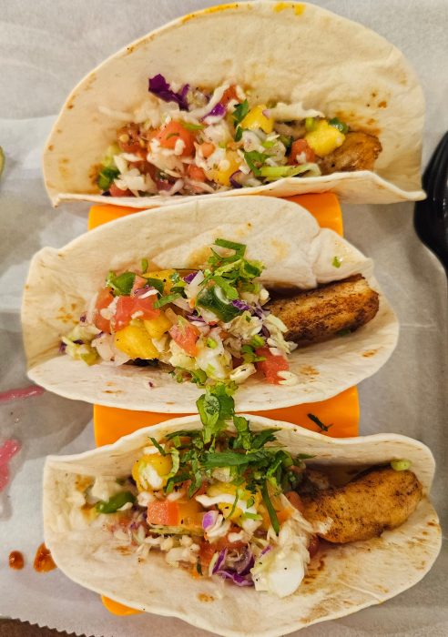Three soft shell tacos filled with seasoned fish, shredded cabbage, diced tomatoes, mango, and chopped cilantro, displayed on an orange taco holder.