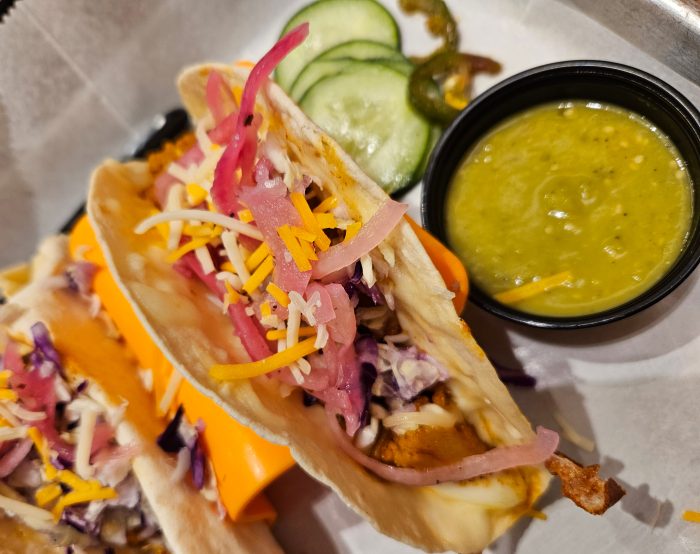 Two soft shell tacos filled with shredded cheese, pickled onions, and slaw, served with sliced cucumbers, jalapeños, and a small cup of green sauce.