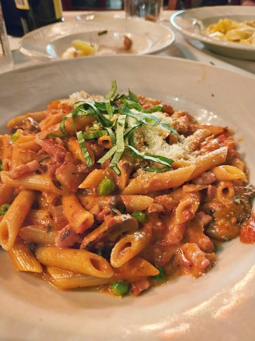 A plate of penne pasta with vegetables, tomato sauce, grated cheese, and fresh basil, served on a white dish with additional plates in the background.