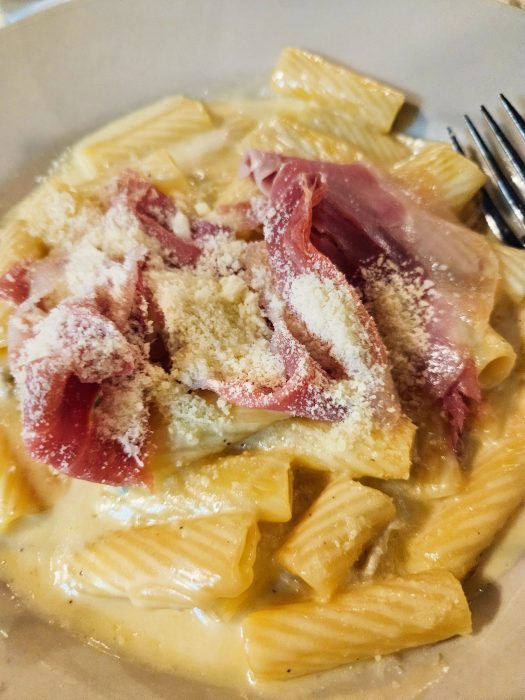 A plate of rigatoni pasta in a creamy sauce, topped with slices of cured meat and grated cheese, with a fork on the side.