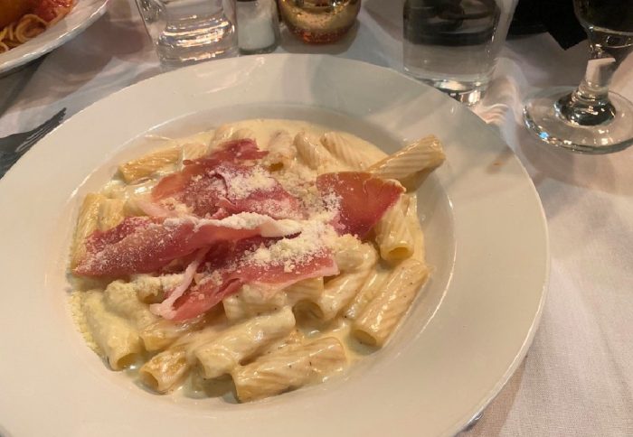 A plate of rigatoni pasta with cream sauce, topped with prosciutto slices and grated cheese, on a white tablecloth with glasses of water and wine nearby.