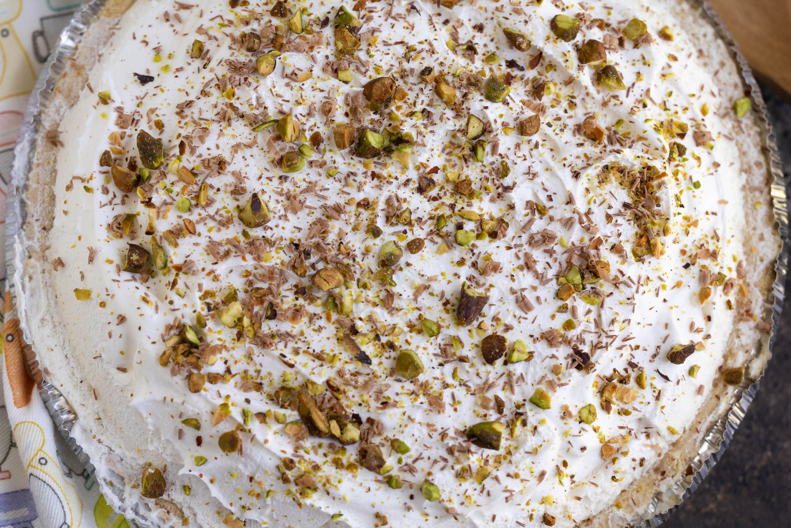 A cream pie topped with chopped pistachios and chocolate shavings in a foil pie dish.