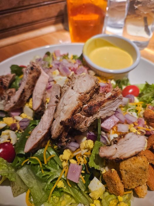 A plate of salad with grilled chicken slices, cherry tomatoes, red onions, shredded cheese, croutons, chopped egg, and a side of yellow dressing. A drink and water glasses are in the background.