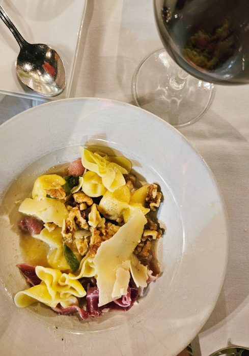 A white plate with pasta, walnuts, shaved cheese, and sauce sits on a table next to a spoon and a glass of red wine.