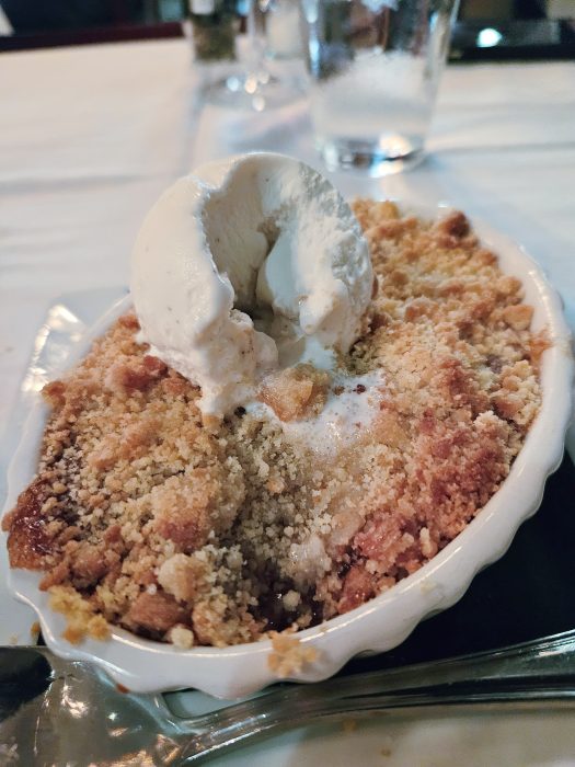 A baked fruit crumble topped with a scoop of vanilla ice cream, served in a white oval dish on a table.