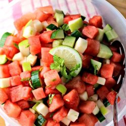 A bowl of diced watermelon, cucumber, and apple salad garnished with mint leaves and a slice of lime, with a spoon on the side.