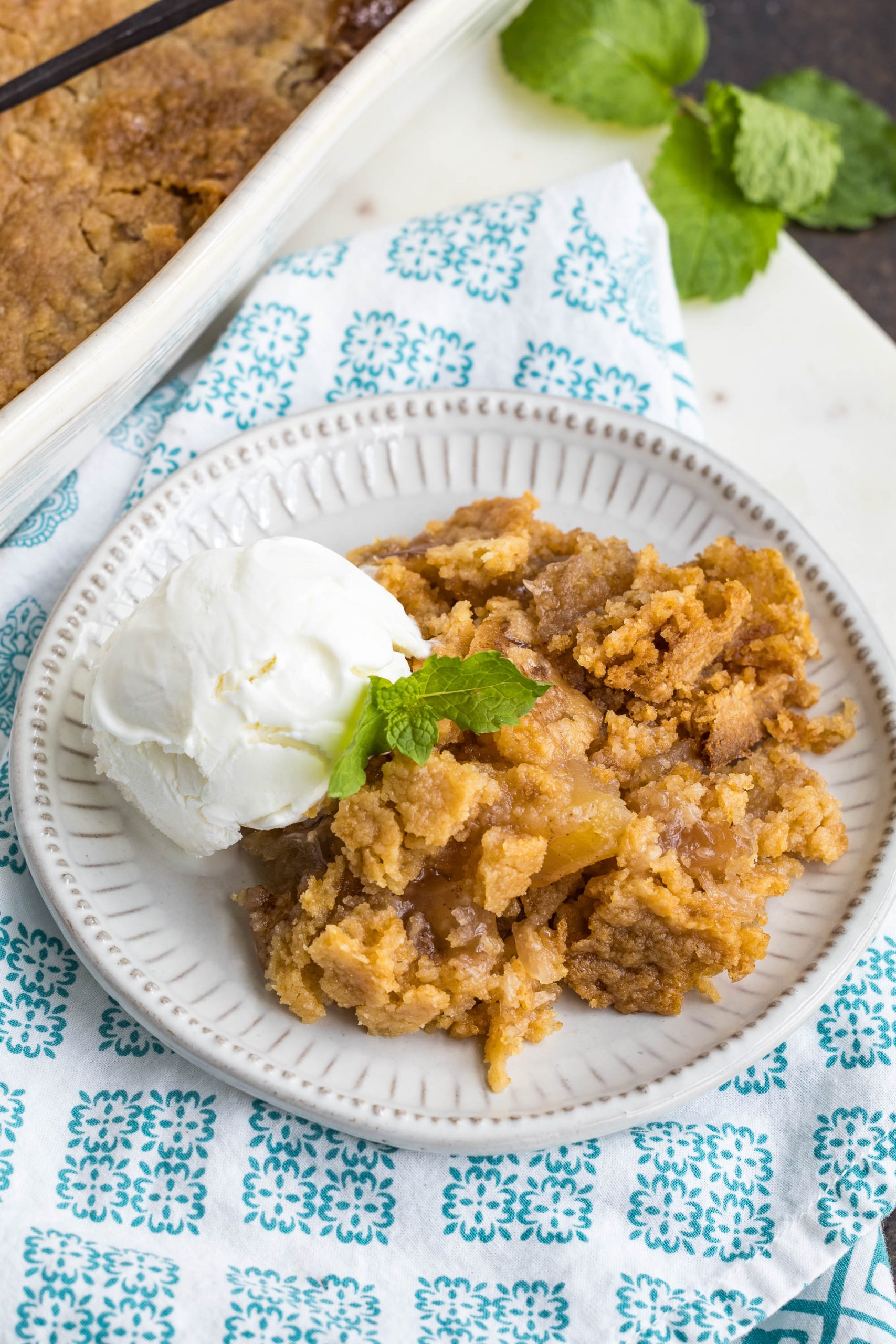 A plate with a serving of apple crisp topped with a sprig of mint, served alongside a scoop of vanilla ice cream on a white dish.