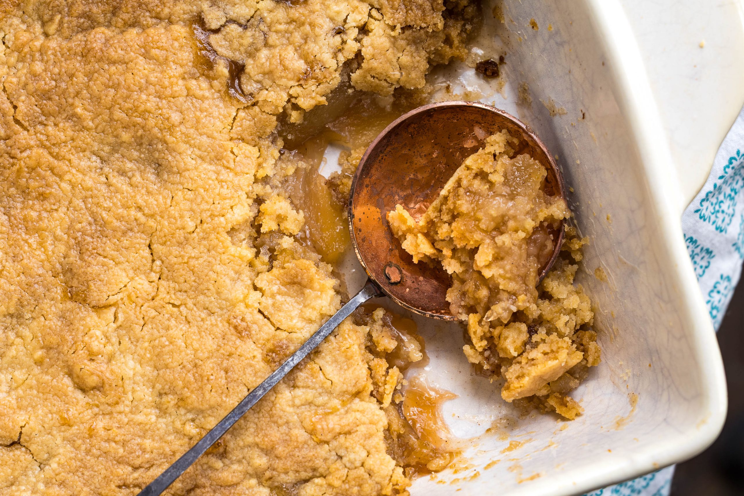A close-up of a partially served cobbler in a white baking dish with a metal serving spoon scooping out a portion.