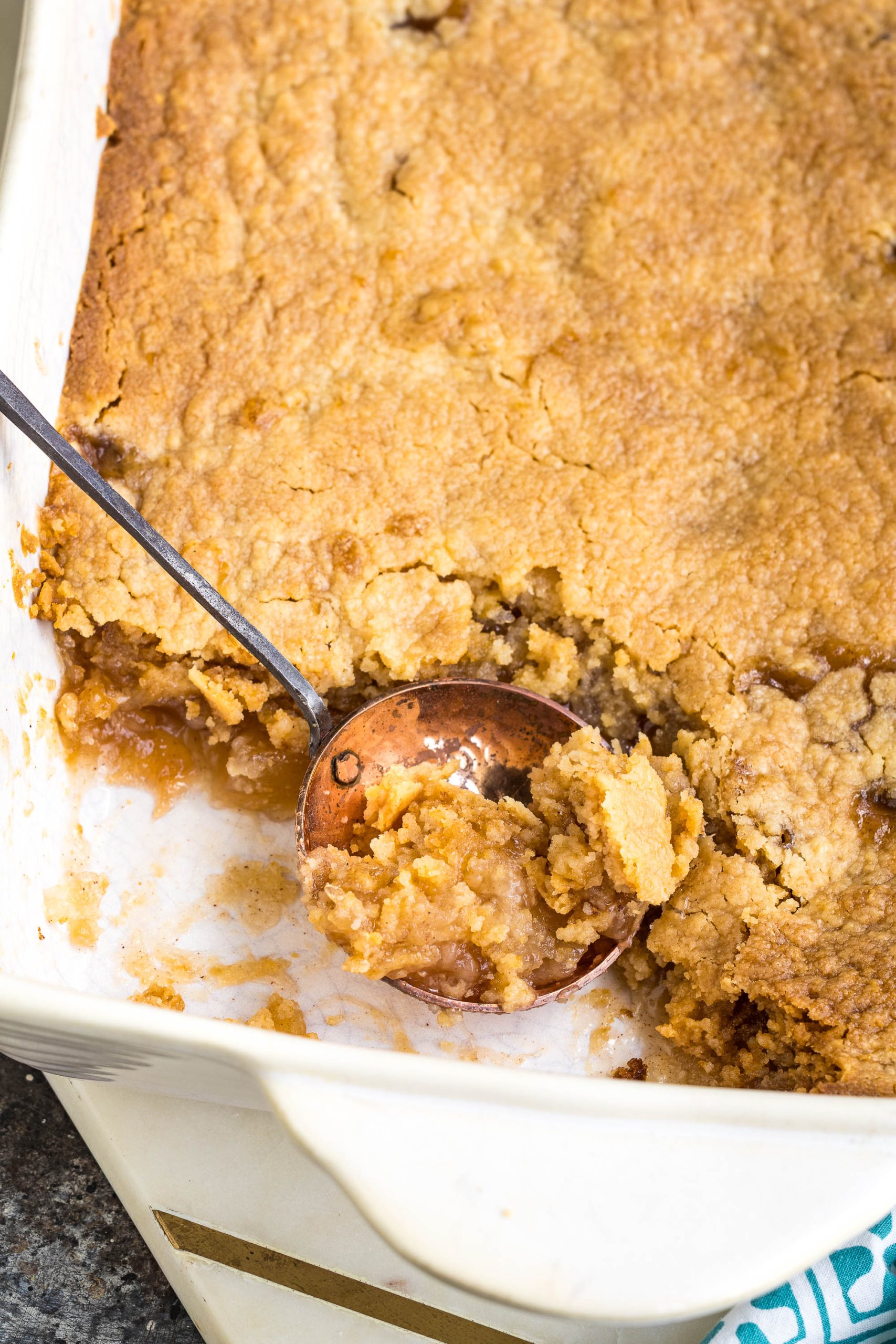 A close-up of a baking dish with a golden-brown cobbler, partially served with a metal spoon revealing its soft, gooey filling underneath the crust.