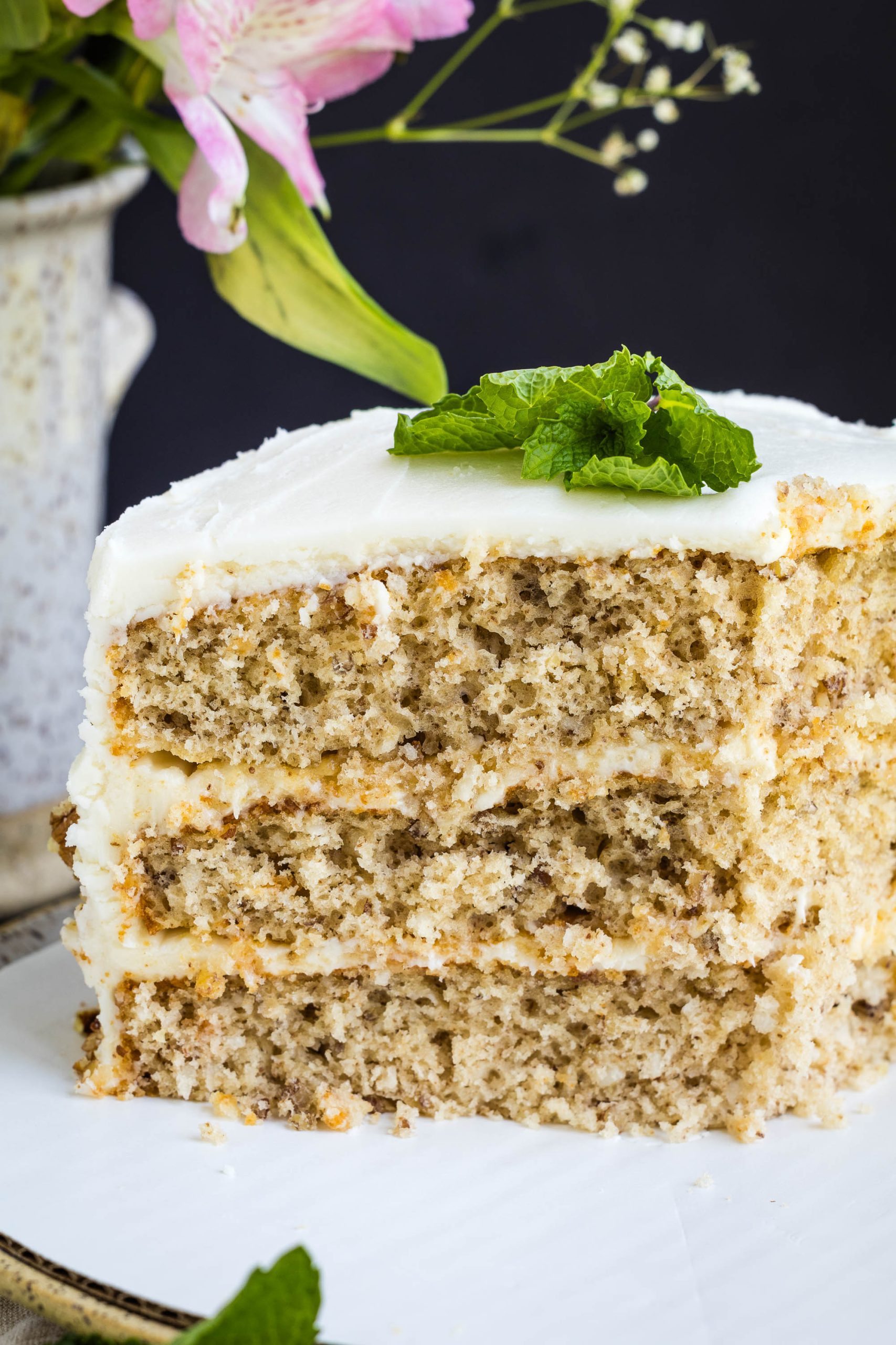 A close-up of a three-layer slice of cake with white frosting, garnished with a sprig of mint, on a white plate.