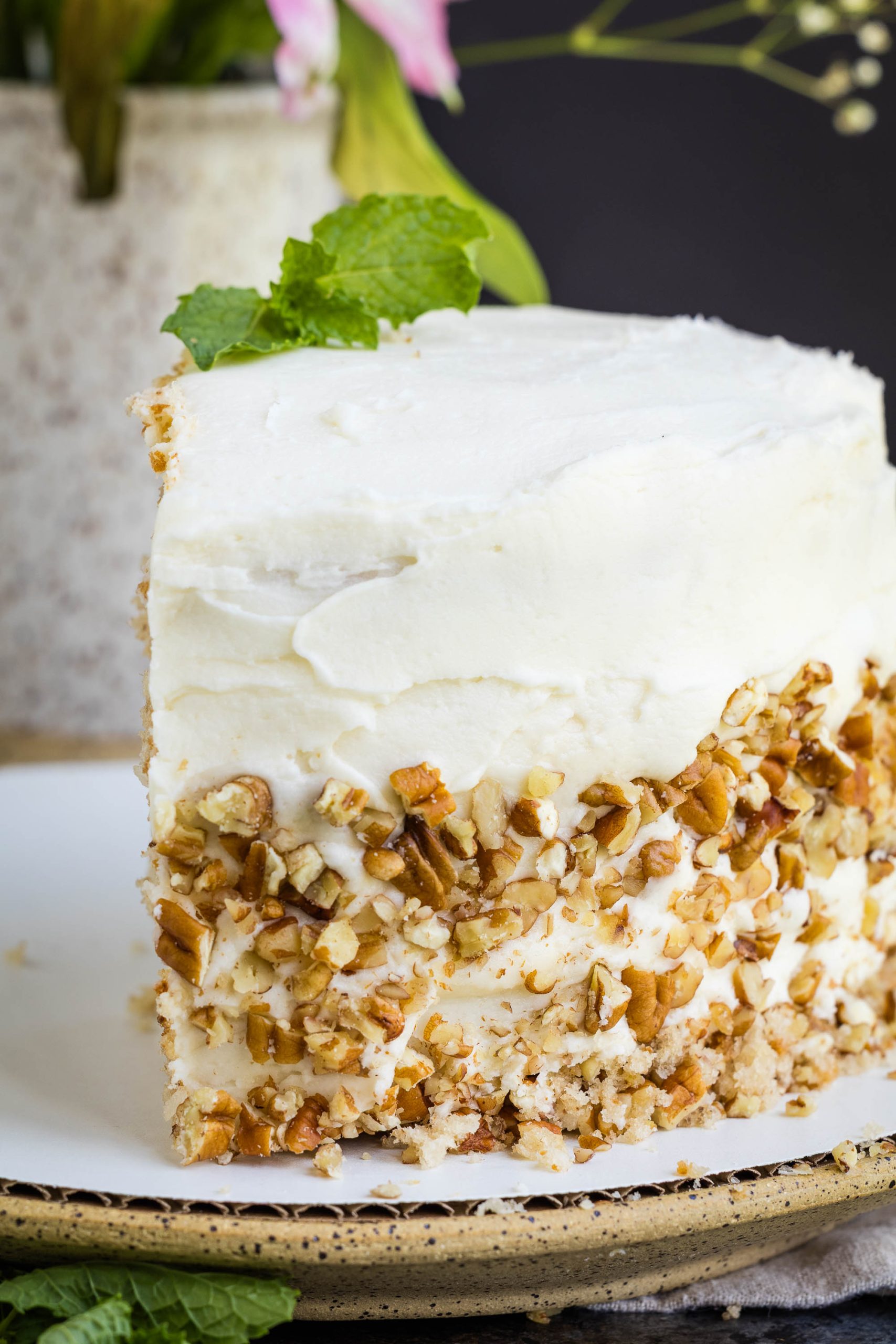A slice of frosted layer cake with chopped pecans on the side sits on a white plate.