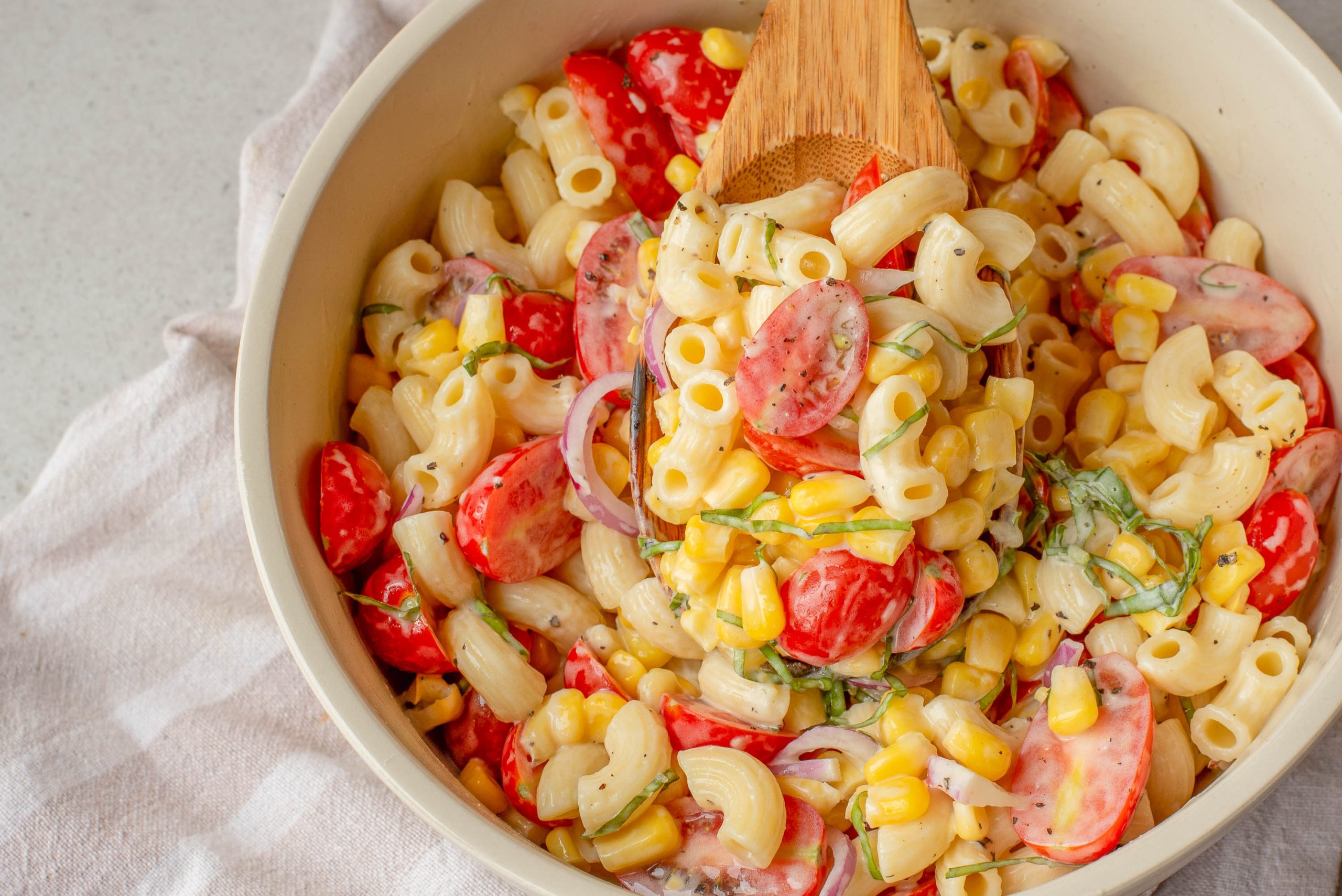 A bowl with macaroni, red onion, fresh herbs, and a creamy dressing, being mixed with a wooden spoon.