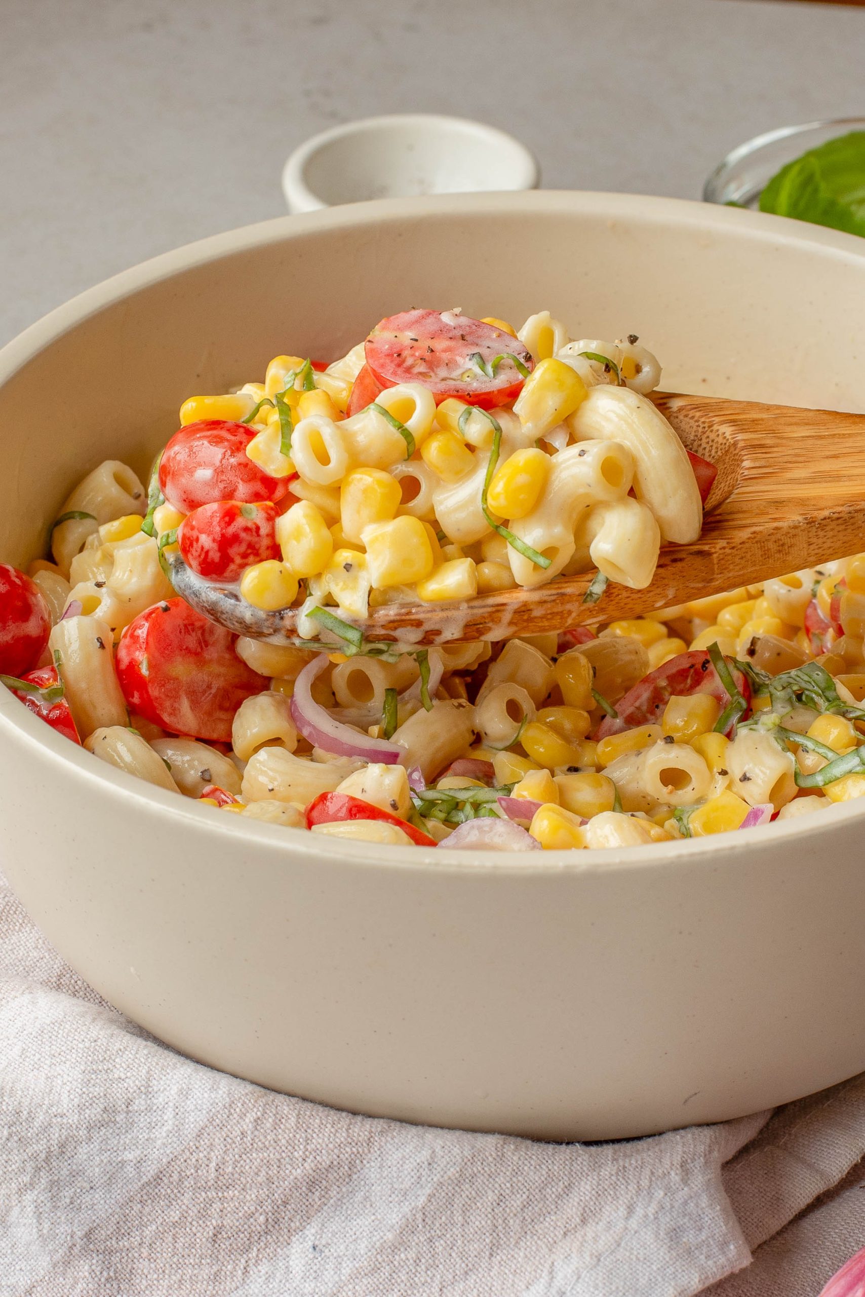 A wooden spoon scoops creamy veggies with corn, cherry tomatoes, red onion, and herbs from a beige bowl.