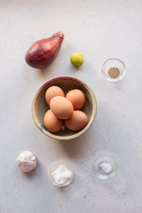 A bowl of brown eggs, an avocado, a lime, a garlic bulb, and small bowls of salt, pepper, and sour cream are arranged on a light countertop.