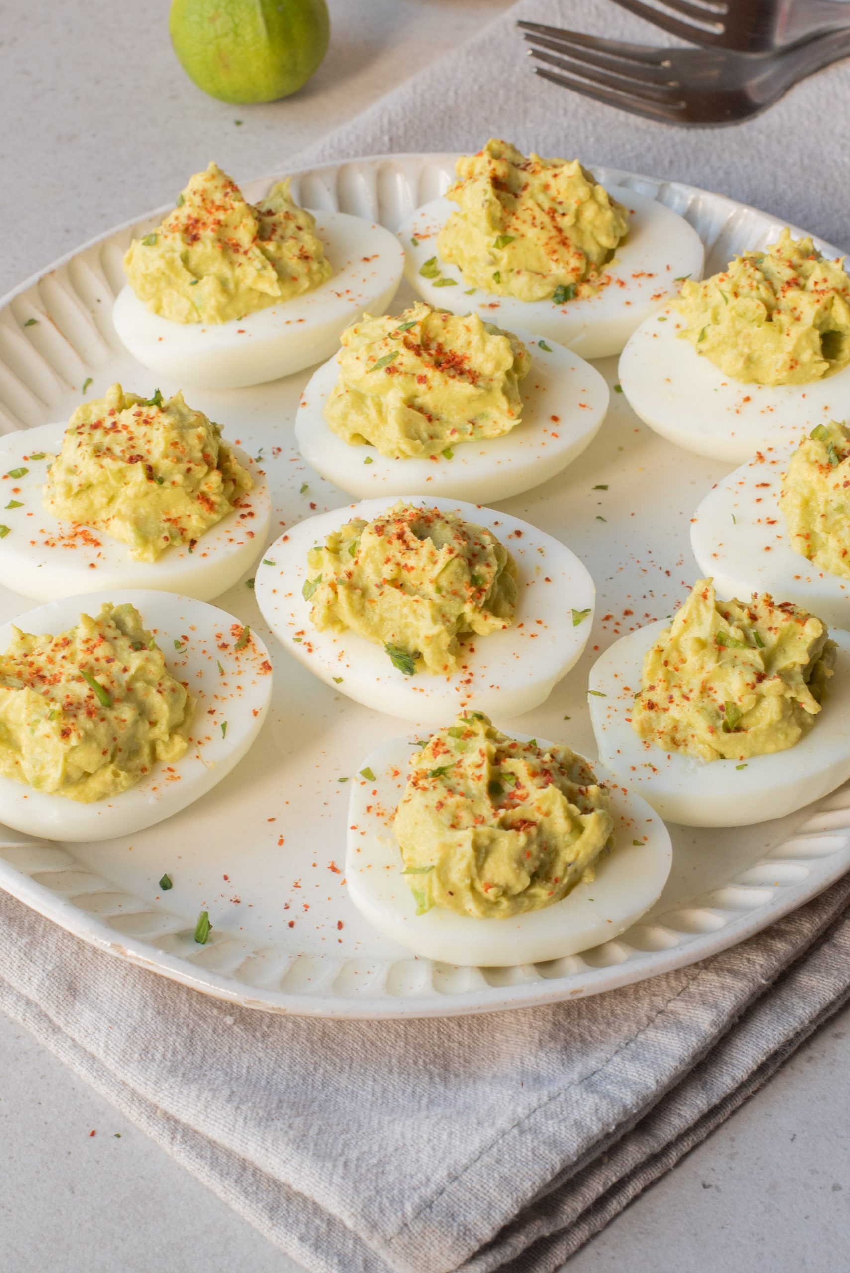A plate of deviled eggs topped with paprika sits on a cloth napkin, with a fork and a lime in the background.