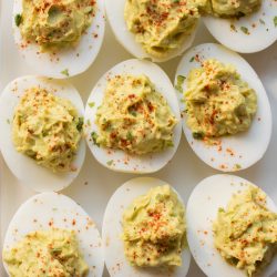 A plate of nine deviled eggs, each topped with a creamy yellow filling and garnished with herbs and paprika.