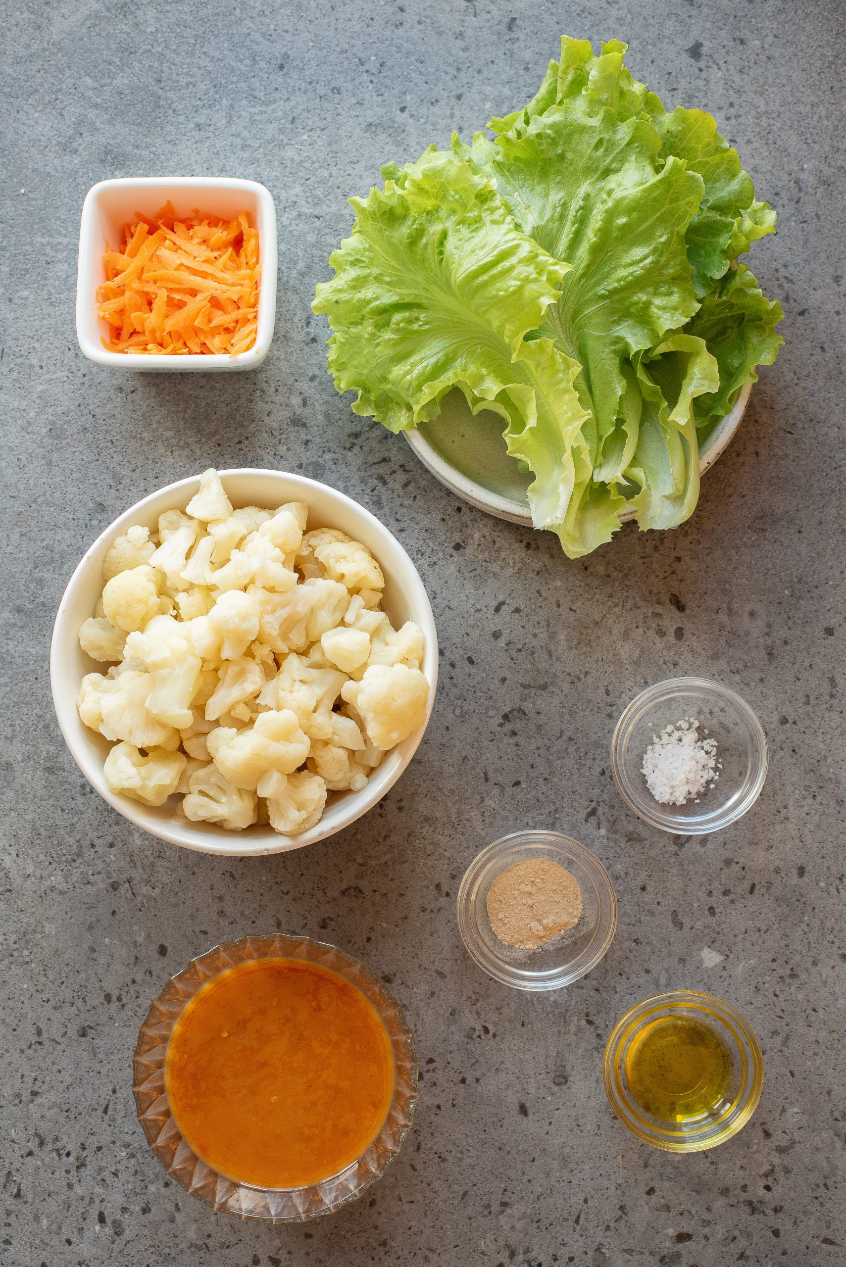 Bowl of chopped cauliflower, lettuce leaves, shredded carrots, salt, ground seasoning, olive oil, and orange dressing arranged on a gray countertop.