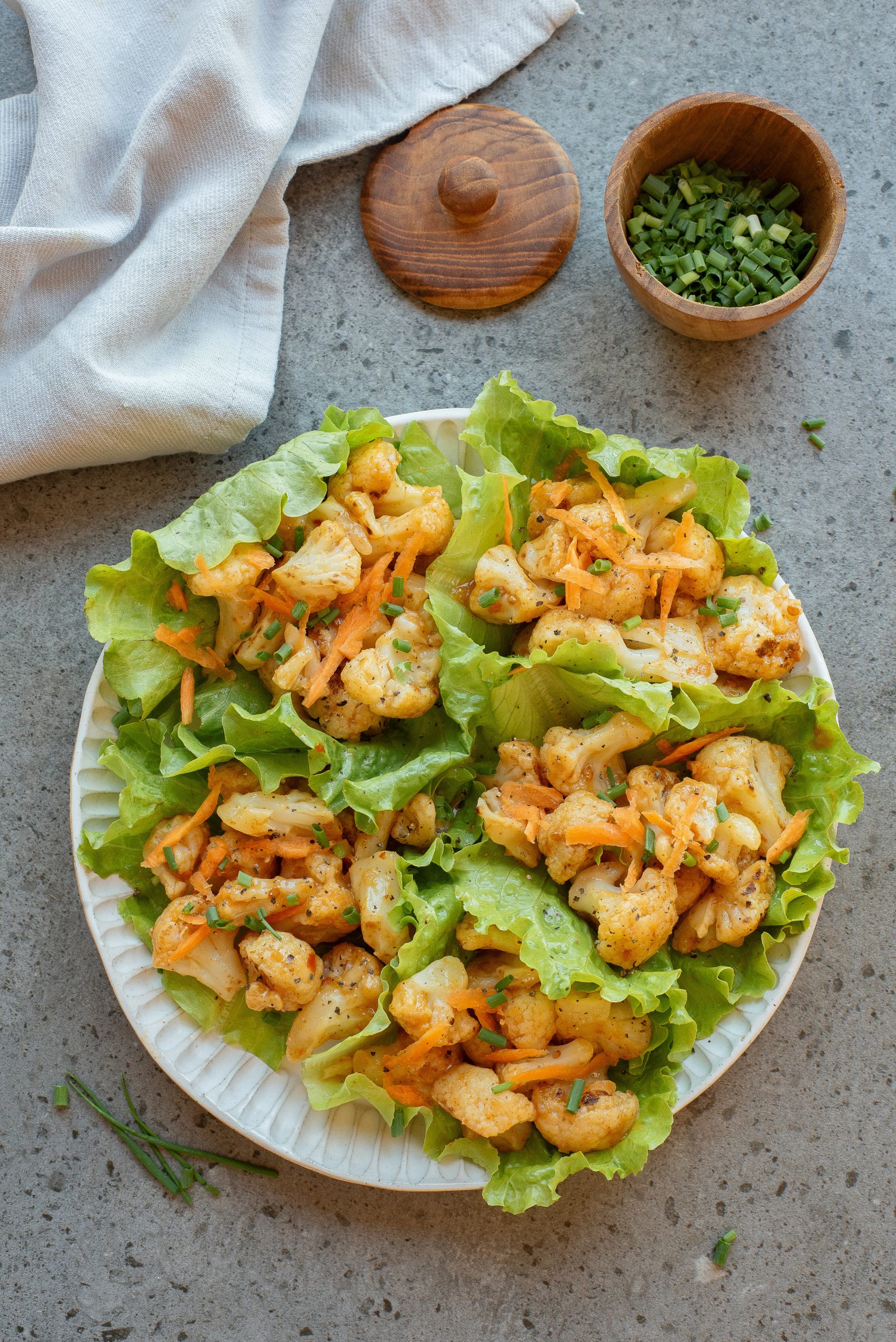 A plate of greens filled with seasoned cauliflower, shredded carrots, and herbs, next to a small bowl of chopped chives on a gray surface.