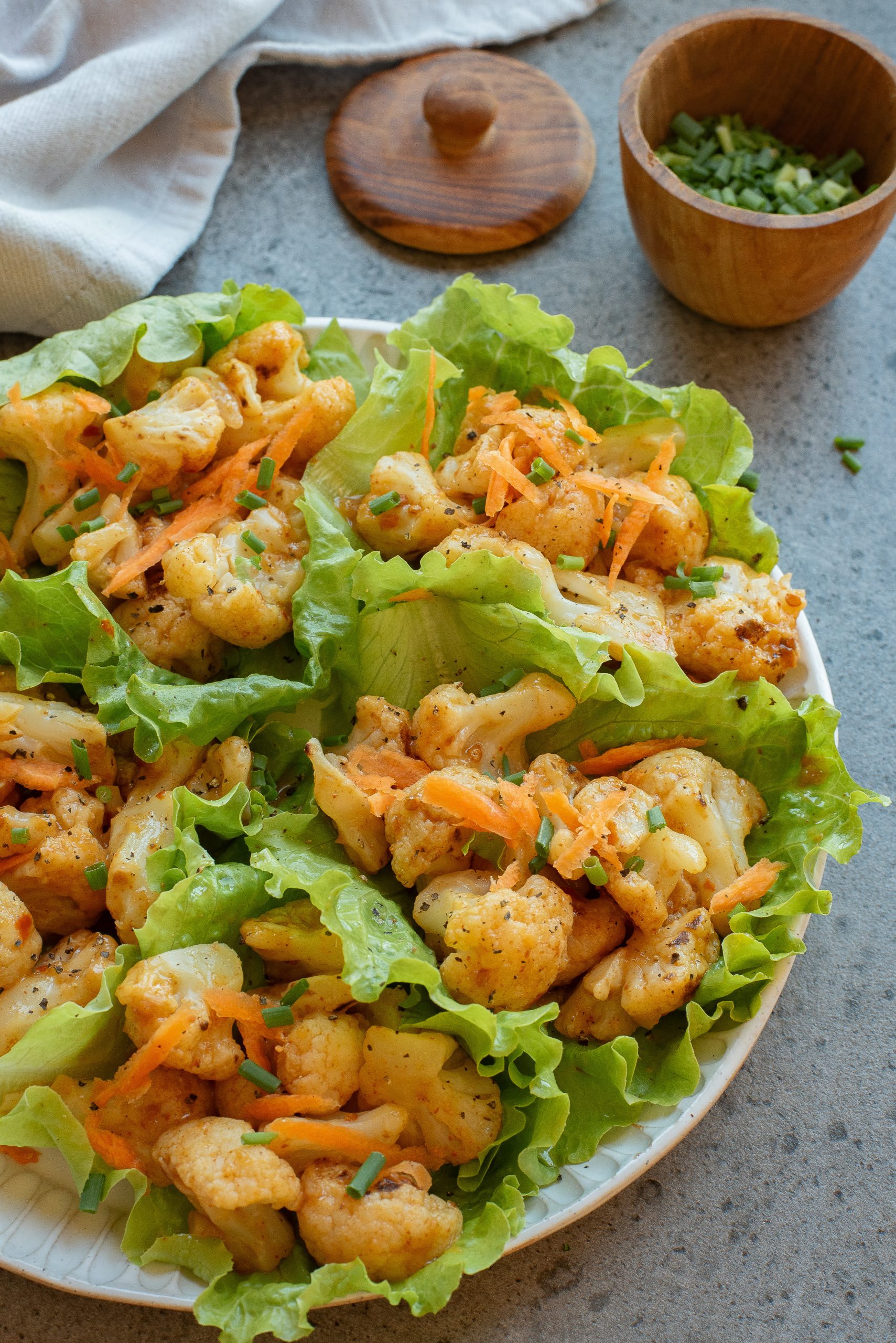 A plate of lettuce filled with seasoned roasted cauliflower and shredded carrots, garnished with chopped chives. A small wooden bowl with more chives is in the background.