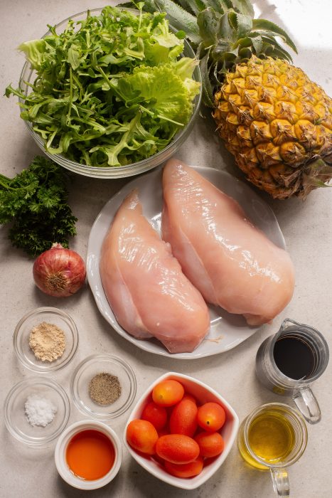 A variety of fresh ingredients on a countertop, including chicken breasts, leafy greens, a pineapple, grape tomatoes, an onion, herbs, and several small bowls of spices and liquids.