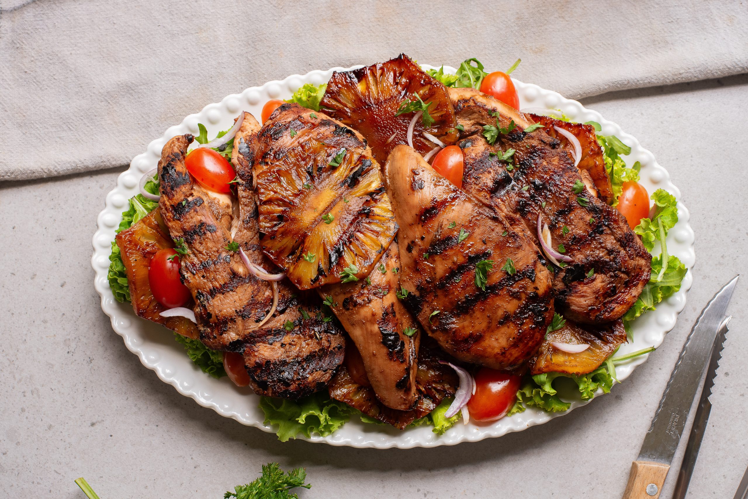 Grilled chicken breasts with charred pineapple slices, cherry tomatoes, and red onion served on a bed of lettuce on a white oval platter.