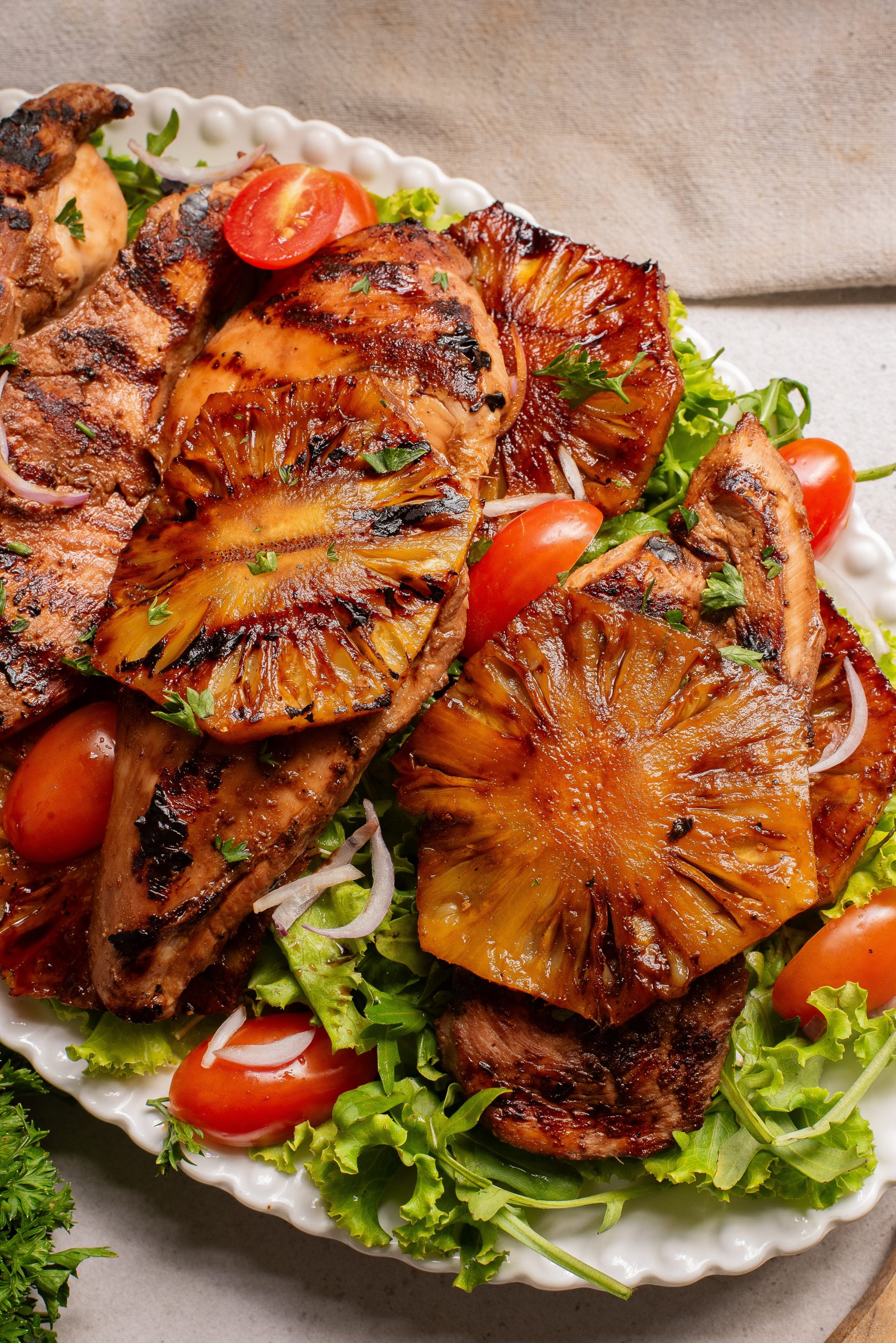 Grilled chicken breasts topped with grilled pineapple slices served on a bed of lettuce, cherry tomatoes, and sliced onions on a white plate.