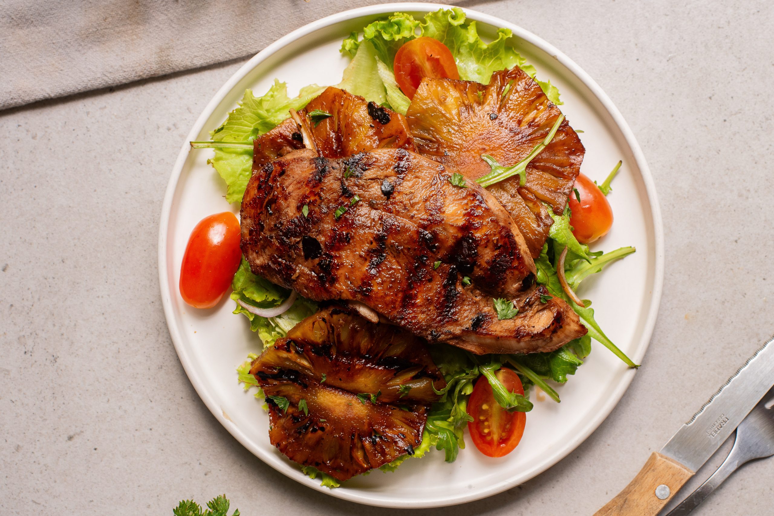 Grilled steak served on a bed of lettuce with grilled pineapple slices and cherry tomatoes on a white plate.