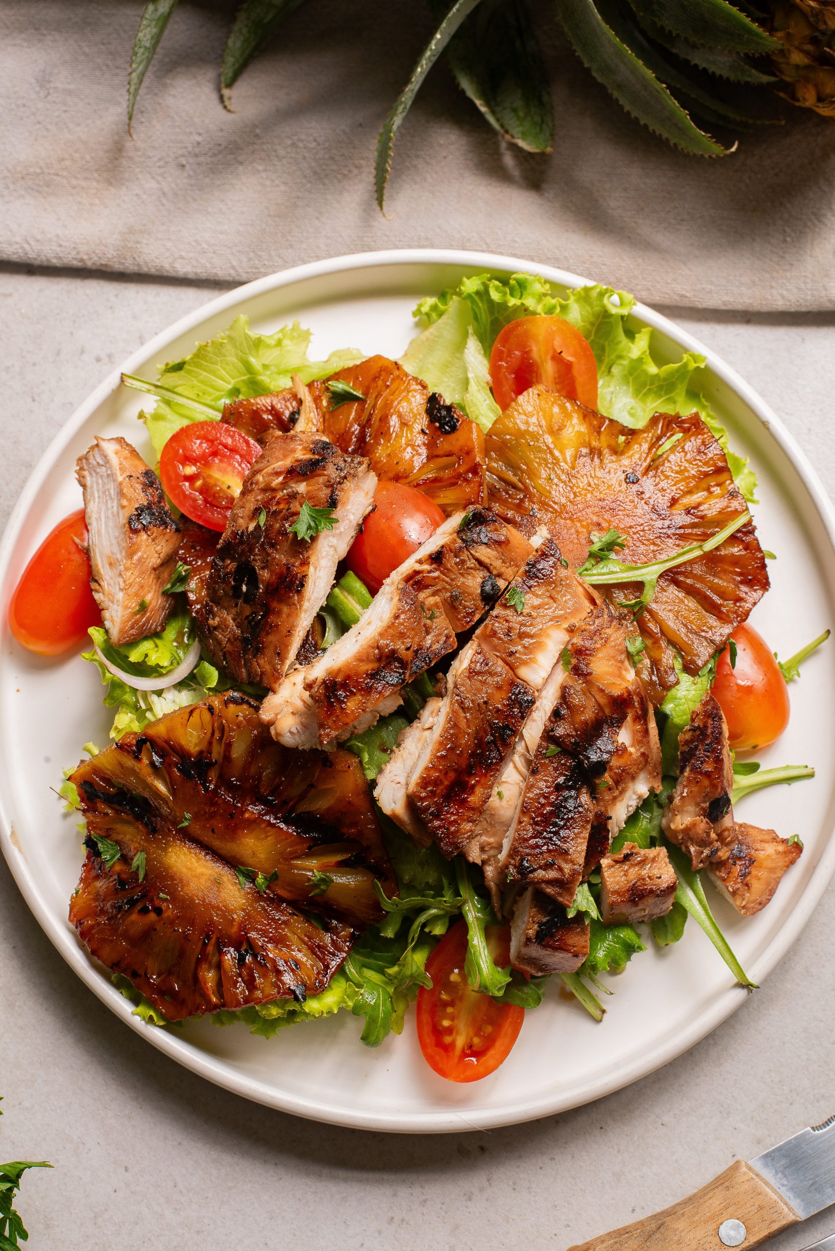 Grilled chicken slices served on a bed of lettuce with grilled pineapple rings and cherry tomato halves on a white plate.