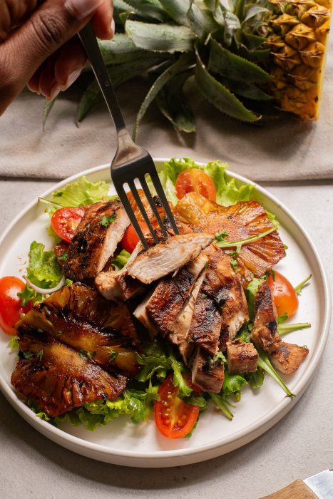 A fork holds a piece of grilled chicken over a plate of sliced chicken, grilled pineapple, lettuce, and cherry tomatoes. A whole pineapple is in the background.