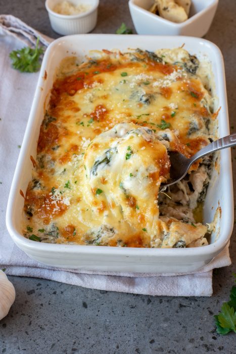 A fork lifting a serving of cheesy baked casserole with spinach from a white rectangular dish on a countertop.