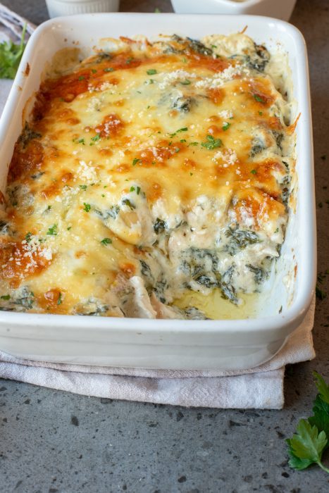 A baked spinach and cheese casserole in a white dish sits on a napkin atop a gray countertop, garnished with chopped herbs.