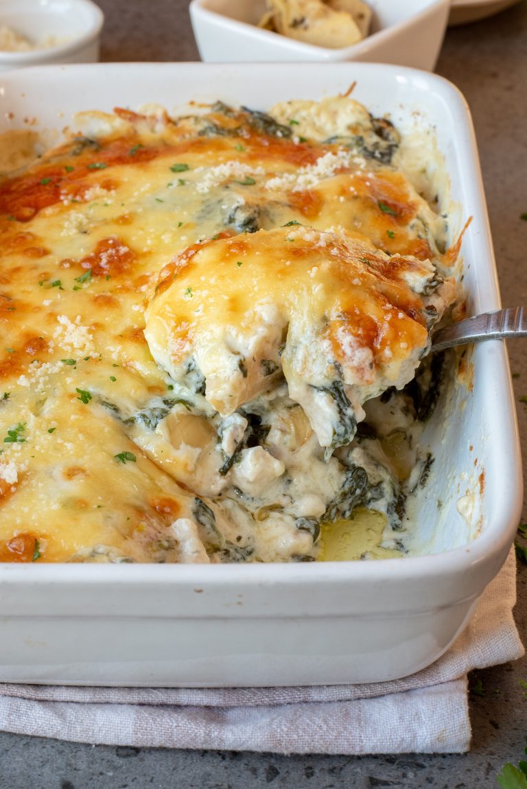 A serving spoon lifts a portion of cheesy spinach and artichoke casserole from a white baking dish.