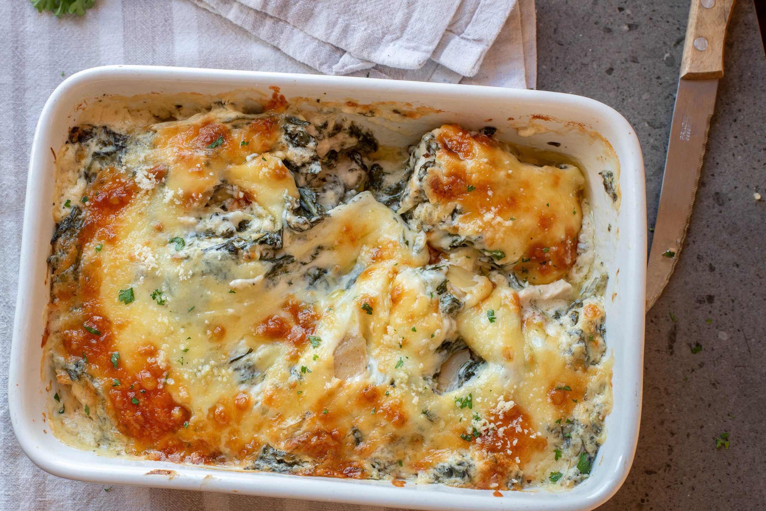 Baked food in a white dish, partially served, with melted golden cheese on top. A knife and white napkin are nearby on a gray surface.