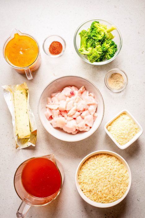 An overhead view of measured ingredients on a countertop, including raw chicken pieces, broccoli, butter, broth, uncooked rice, grated cheese, seasonings, and a sauce.