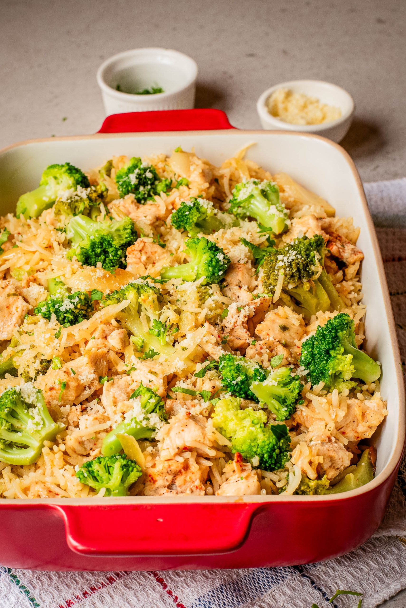 A red baking dish filled with a rice casserole containing pieces of chicken, broccoli florets, and shredded cheese, with small bowls in the background.