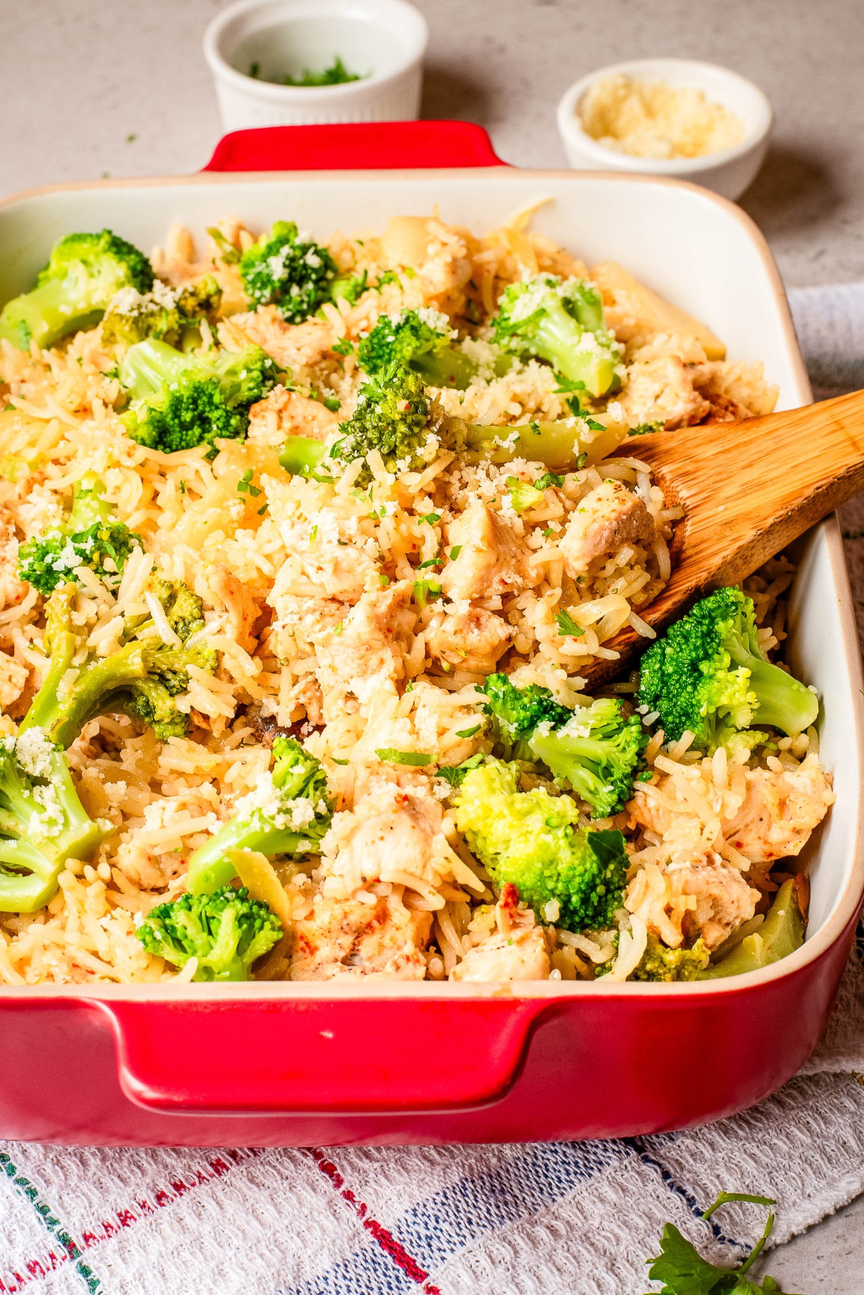 A casserole dish filled with cooked rice, broccoli florets, and pieces of chicken, being stirred with a wooden spoon. Small bowls with grated cheese and parsley are in the background.