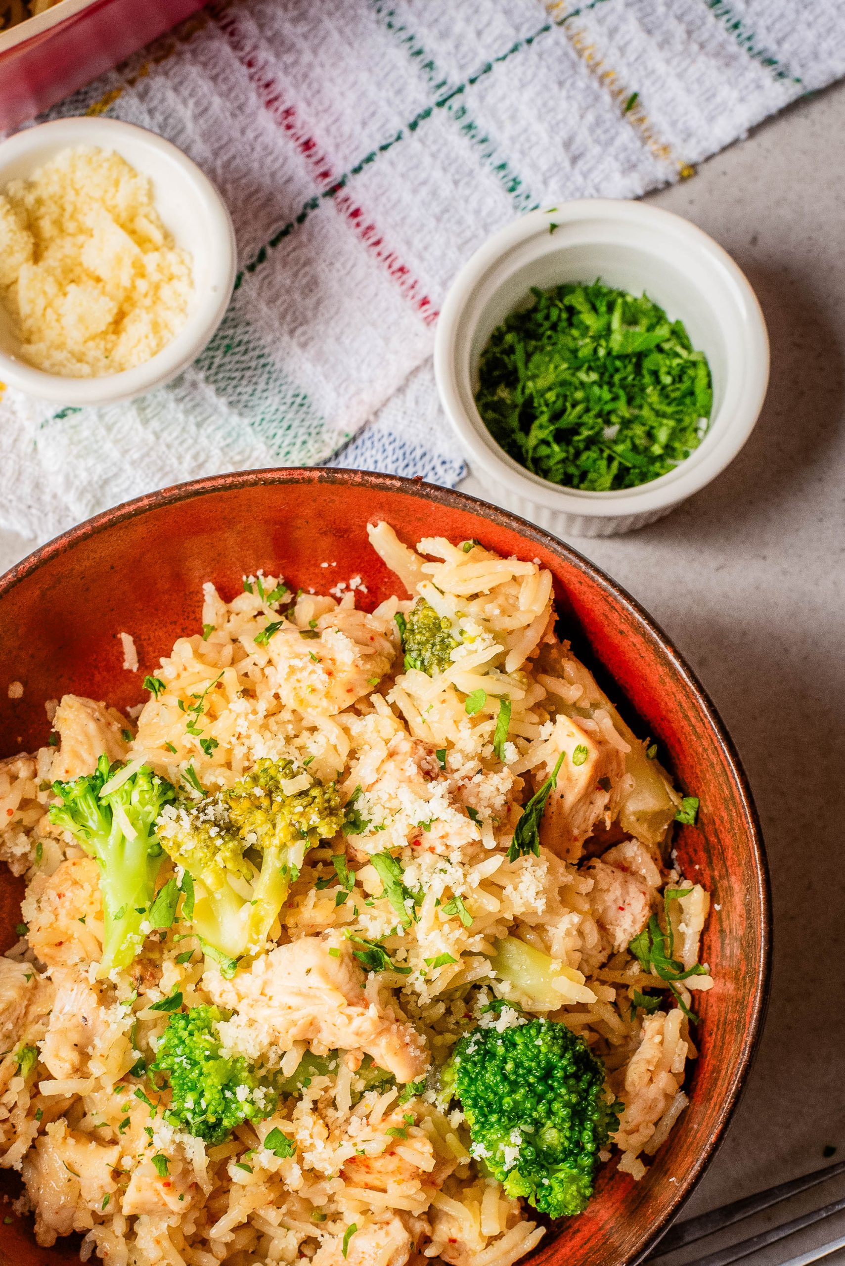 A bowl of rice mixed with broccoli and chicken is on a table, with small bowls of grated cheese and chopped herbs nearby.