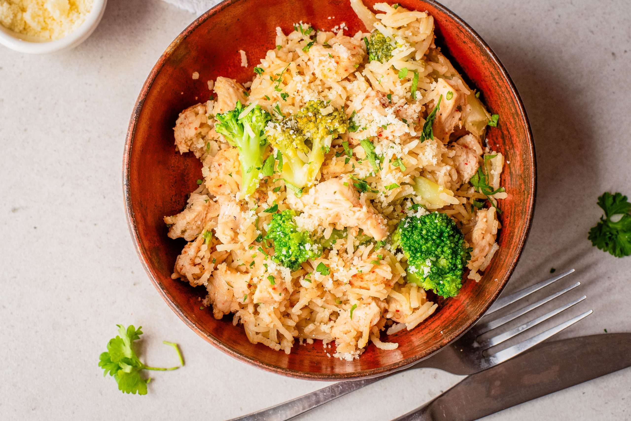 A bowl of rice mixed with pieces of chicken and broccoli, garnished with herbs, placed on a light surface next to a fork and parsley.