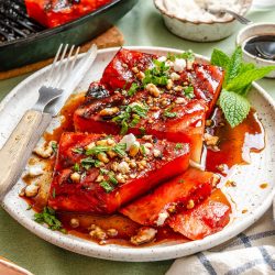 Grilled watermelon slices topped with crumbled cheese, chopped herbs, nuts, and a drizzle of balsamic glaze, served on a white plate with a fork and mint garnish.