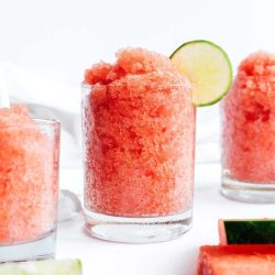 Three glasses filled with red, icy, slushy drinks, each garnished with a lime slice, with watermelon slices nearby on a white surface.