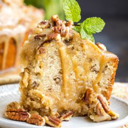 A slice of pecan bundt cake with glaze, garnished with pecans and a sprig of mint, served on a patterned plate.