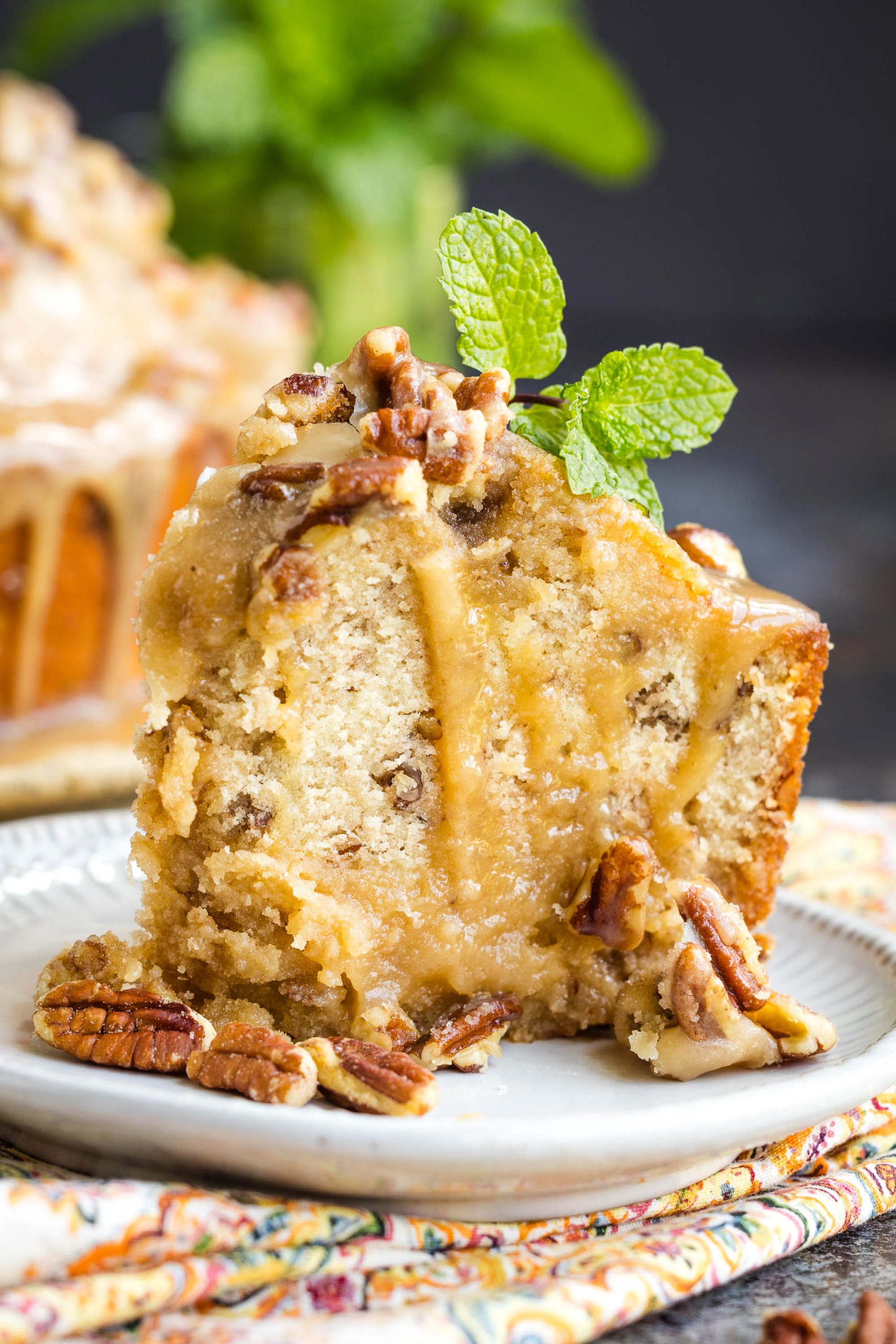 A slice of Bourbon Praline Pecan Pound Cake with glaze, garnished with pecans and a sprig of mint, served on a patterned plate.