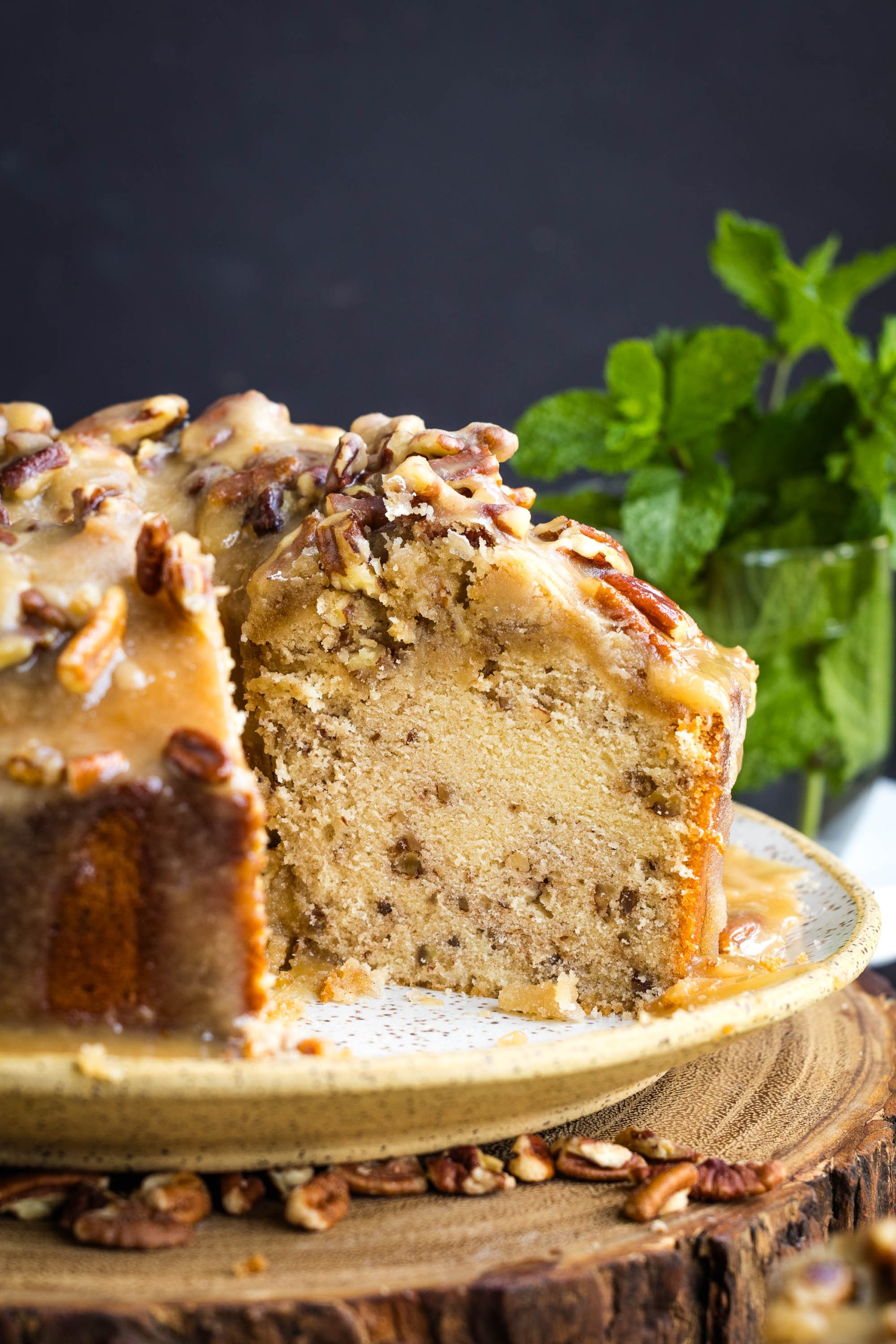 A sliced bundt cake with a caramel glaze and chopped pecans on top, displayed on a plate with fresh mint in the background.