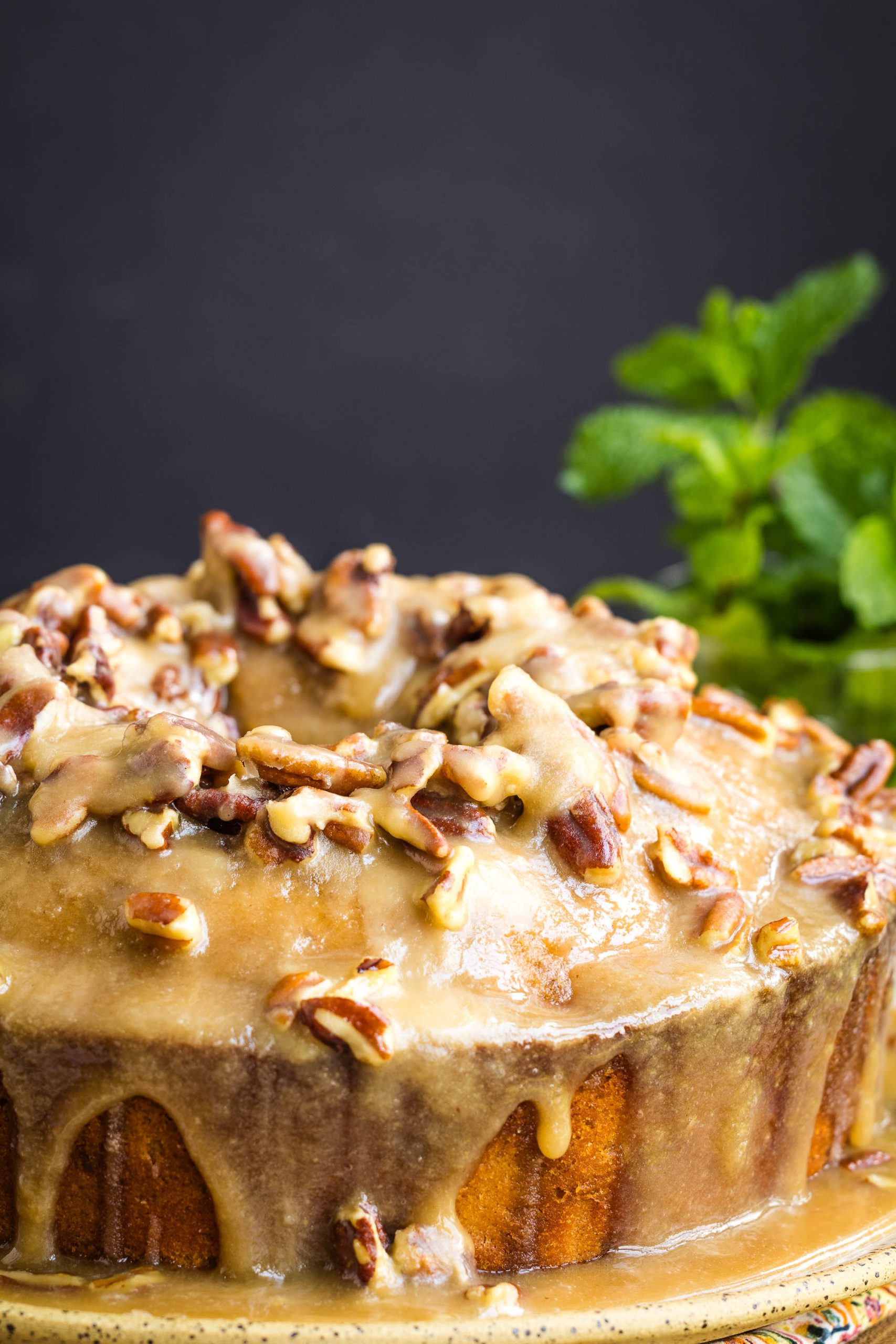 A round cake topped with caramel glaze and chopped pecans, with glaze dripping down the sides; a sprig of mint is in the background.