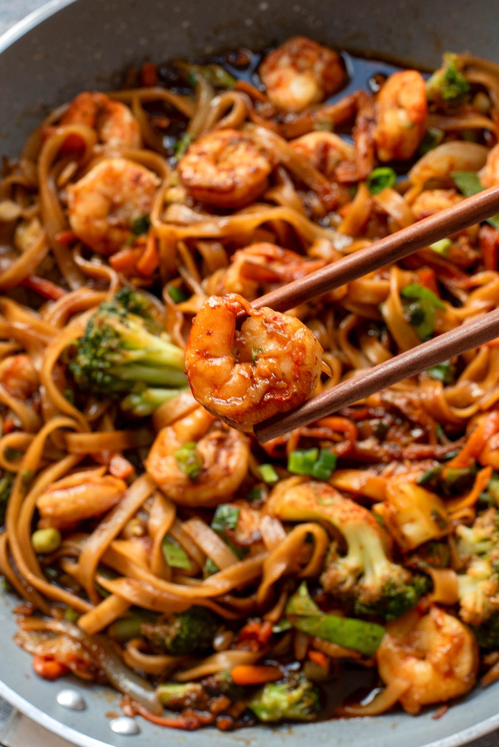 Chopsticks holding a shrimp above a pan of stir-fried noodles with shrimp, broccoli, and vegetables.