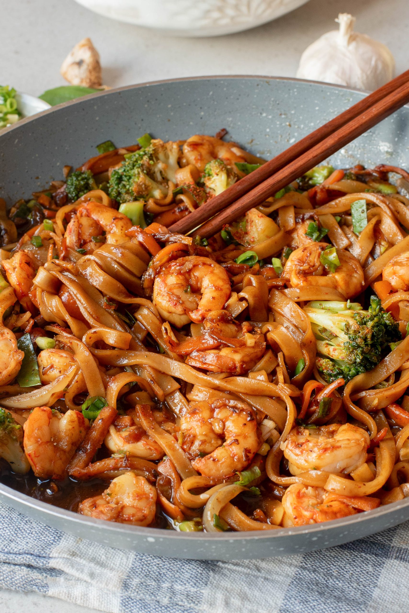 A pan filled with shrimp stir-fried drunken Thai noodles, broccoli, and vegetables, with chopsticks picking up some noodles.
