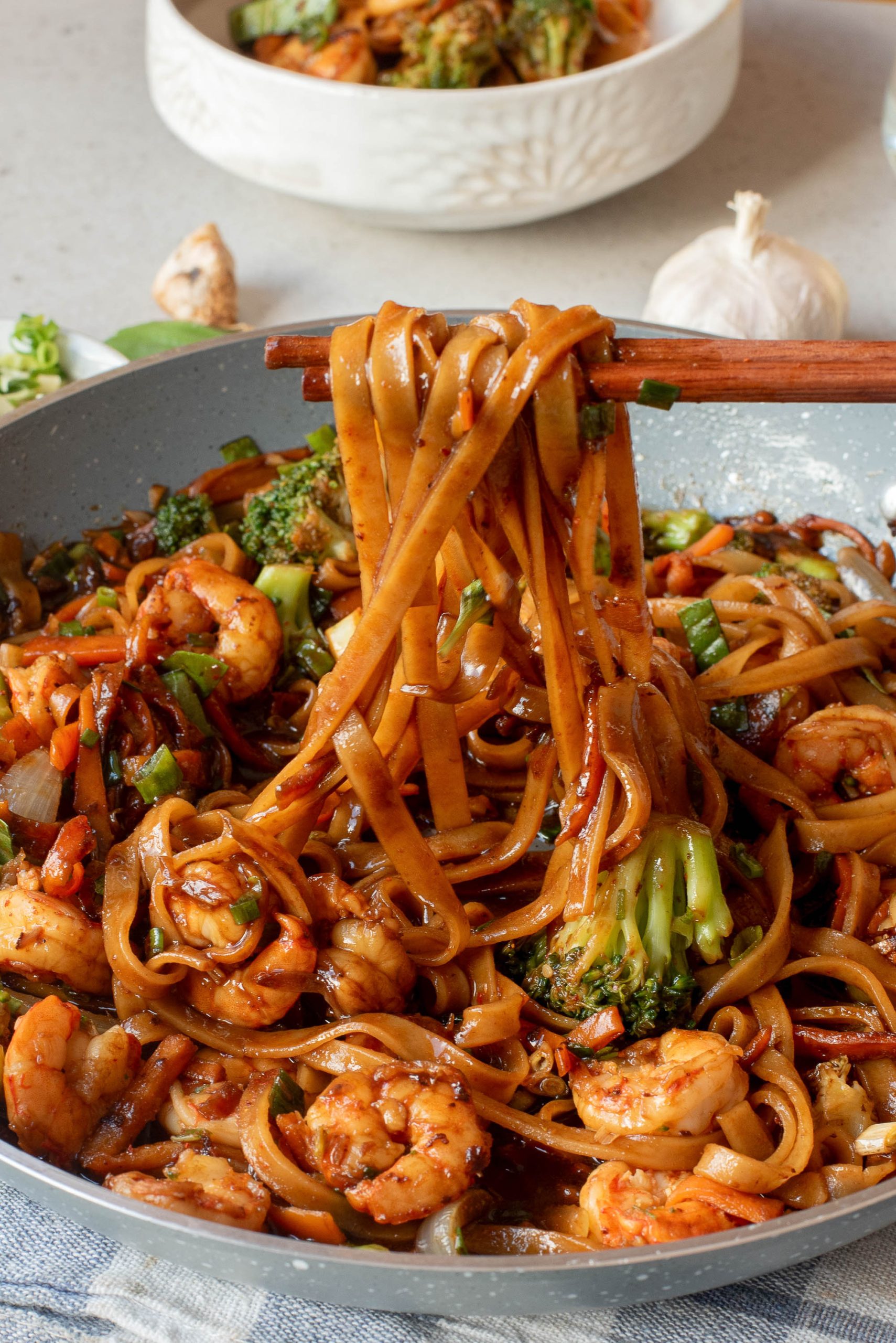 A pan of drunken Thai noodles with shrimp, broccoli, and vegetables is being lifted with chopsticks.